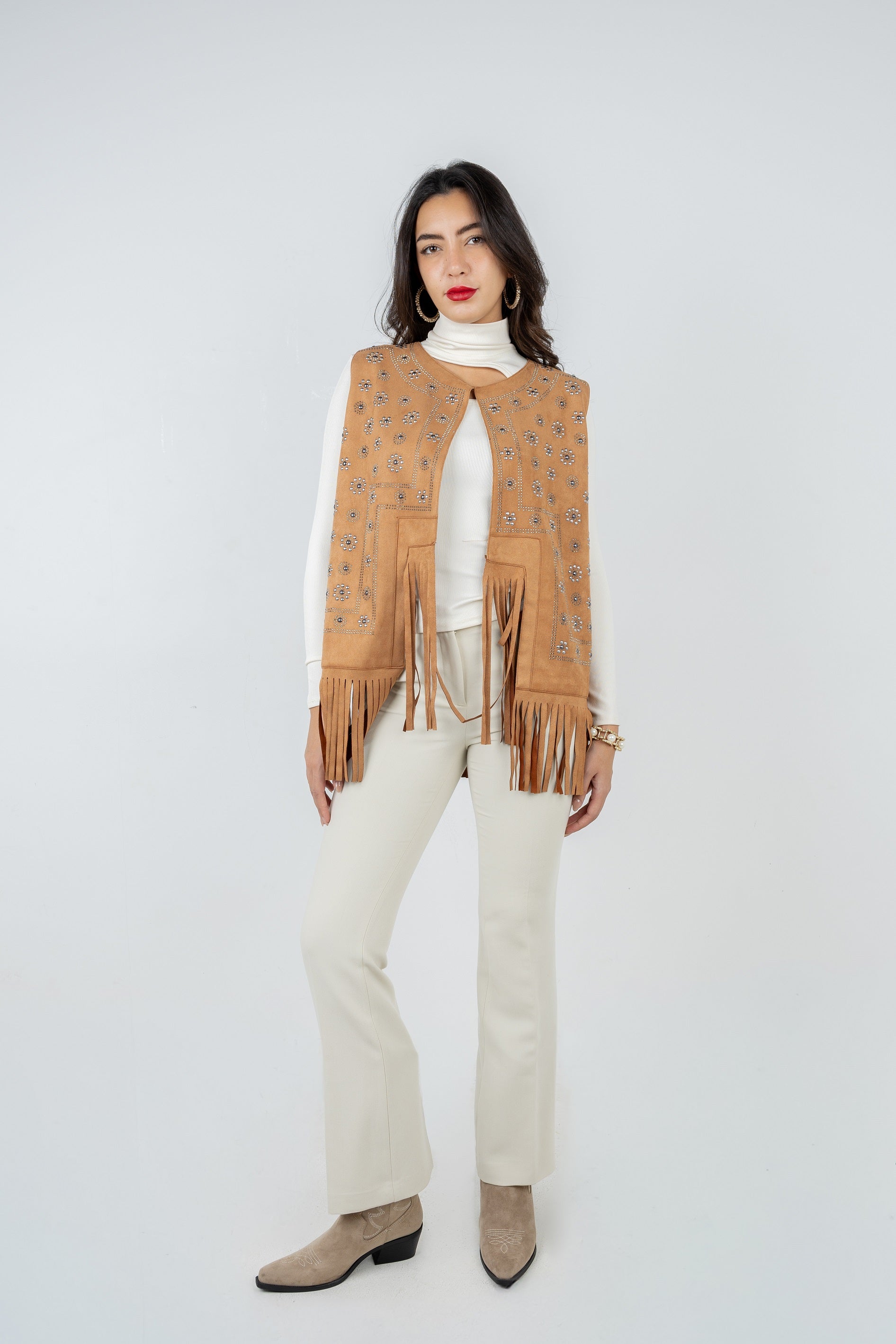 Woman wearing a tan fringe vest over a white turtleneck and pants on a white background