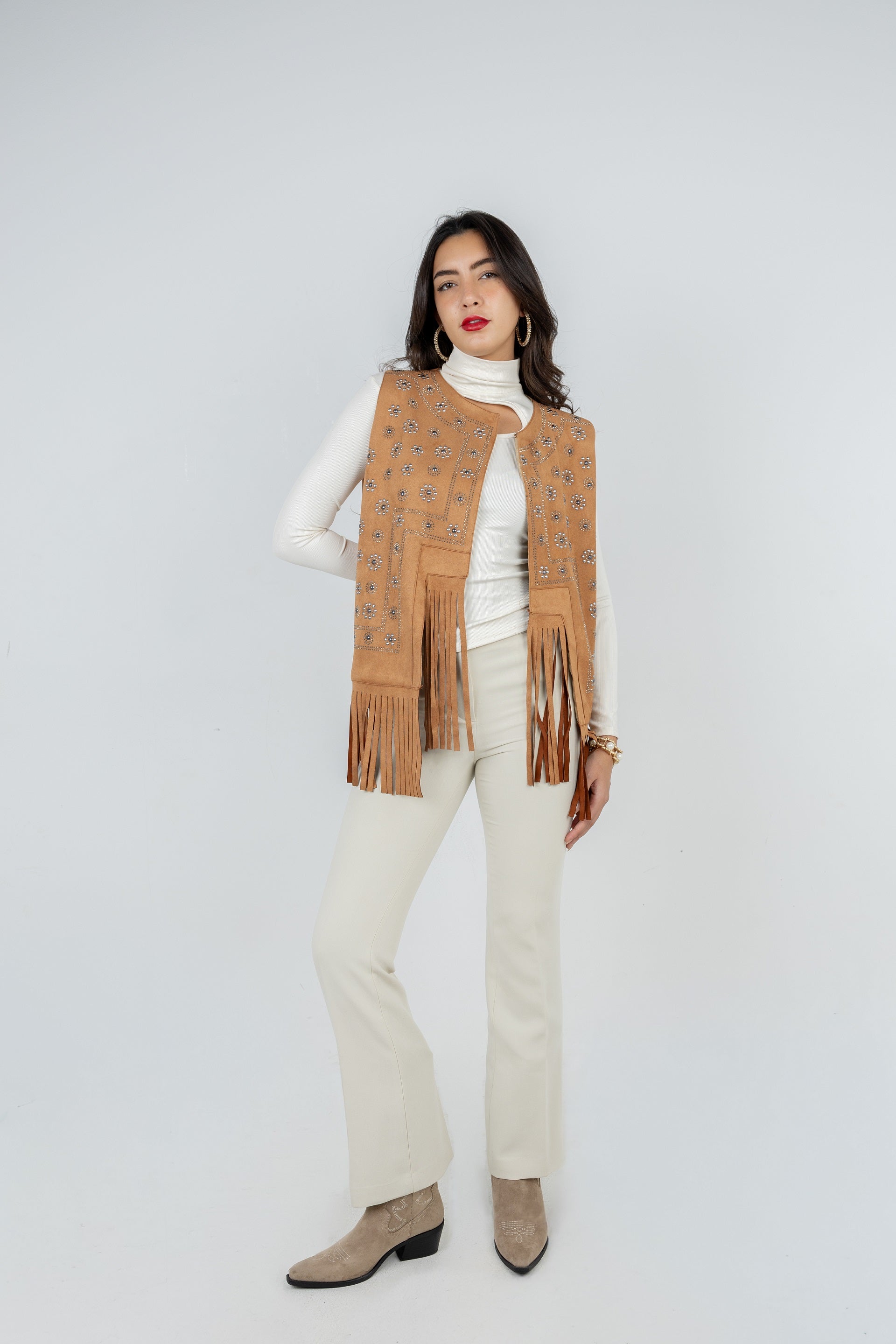 Woman wearing a beige fringed vest over a white turtleneck and pants on a white background