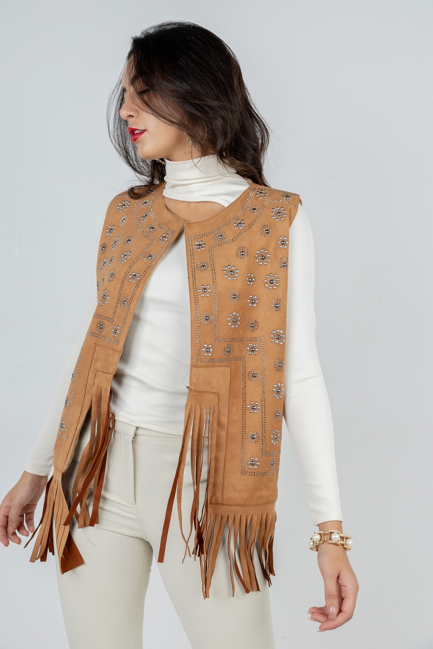 Woman wearing a tan fringed vest over a white long-sleeve shirt on a light gray background