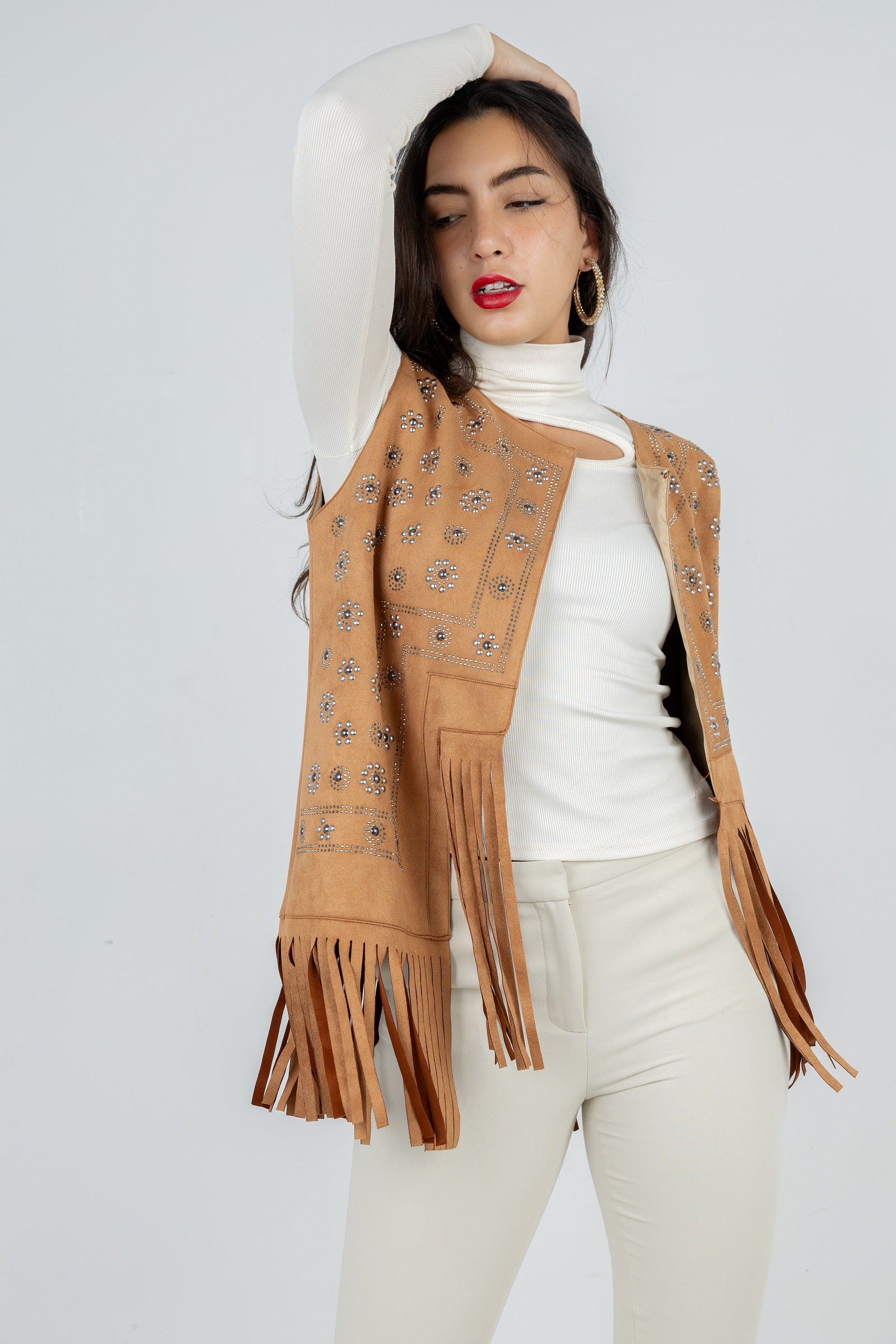Woman wearing a tan fringed vest over a white turtleneck and pants on a light gray background