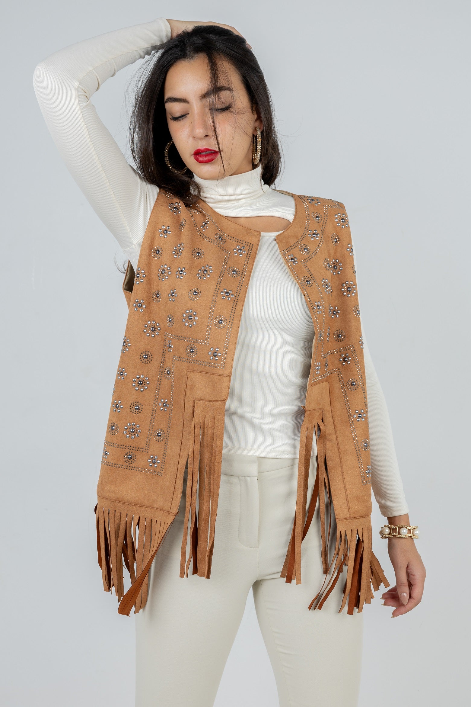 Woman wearing a tan fringed vest over a white turtleneck and pants on a light gray background