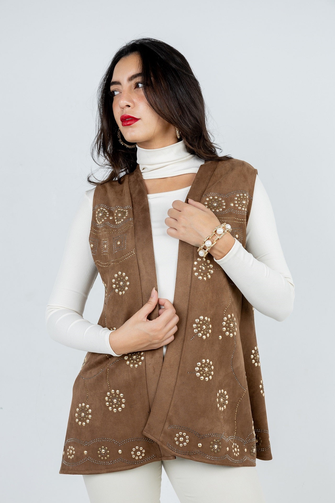 Woman wearing a brown embroidered vest over a white long-sleeve shirt on a light gray background