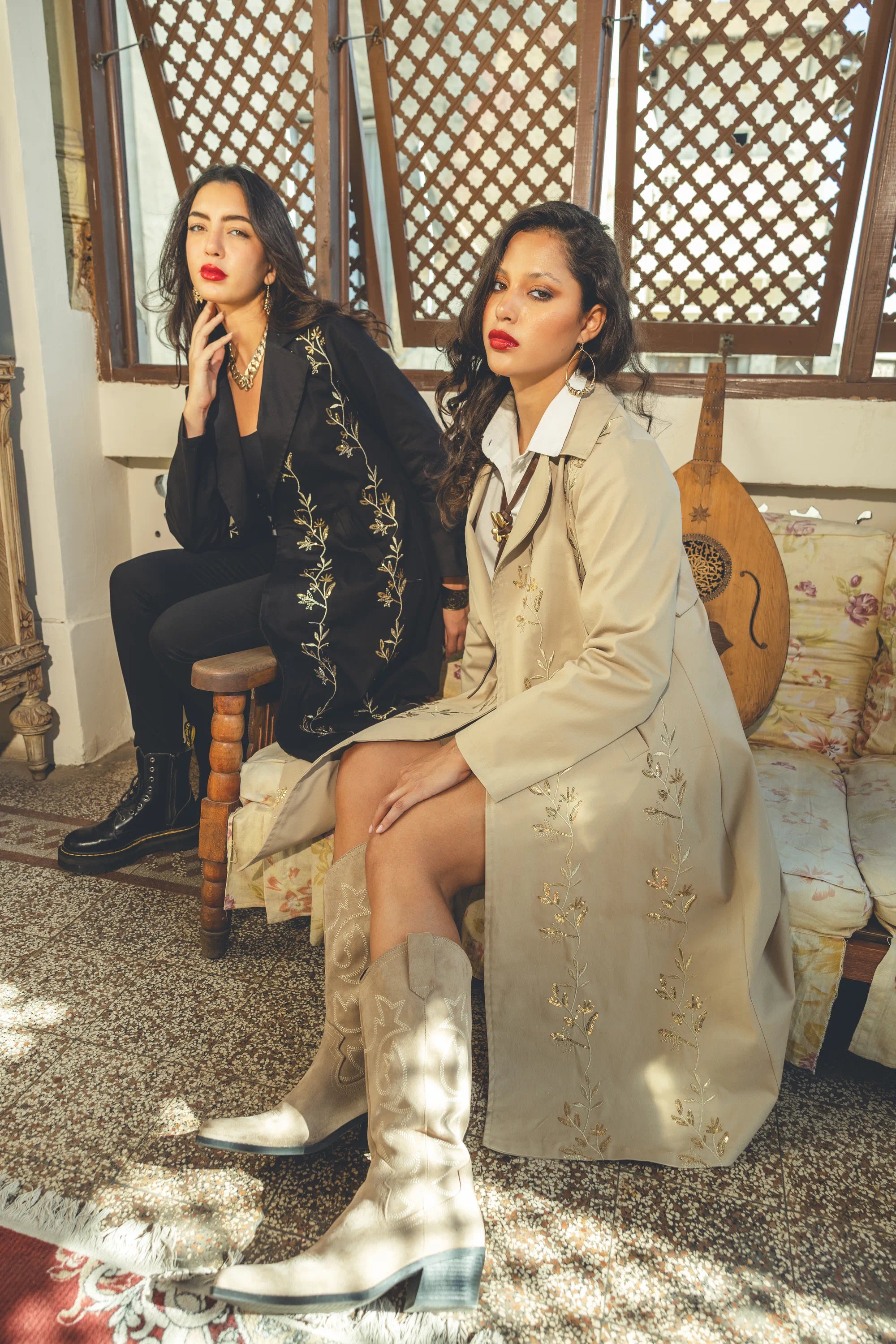 Two women sitting in a decorative interior setting, one in a black outfit and the other in a beige coat with floral patterns.