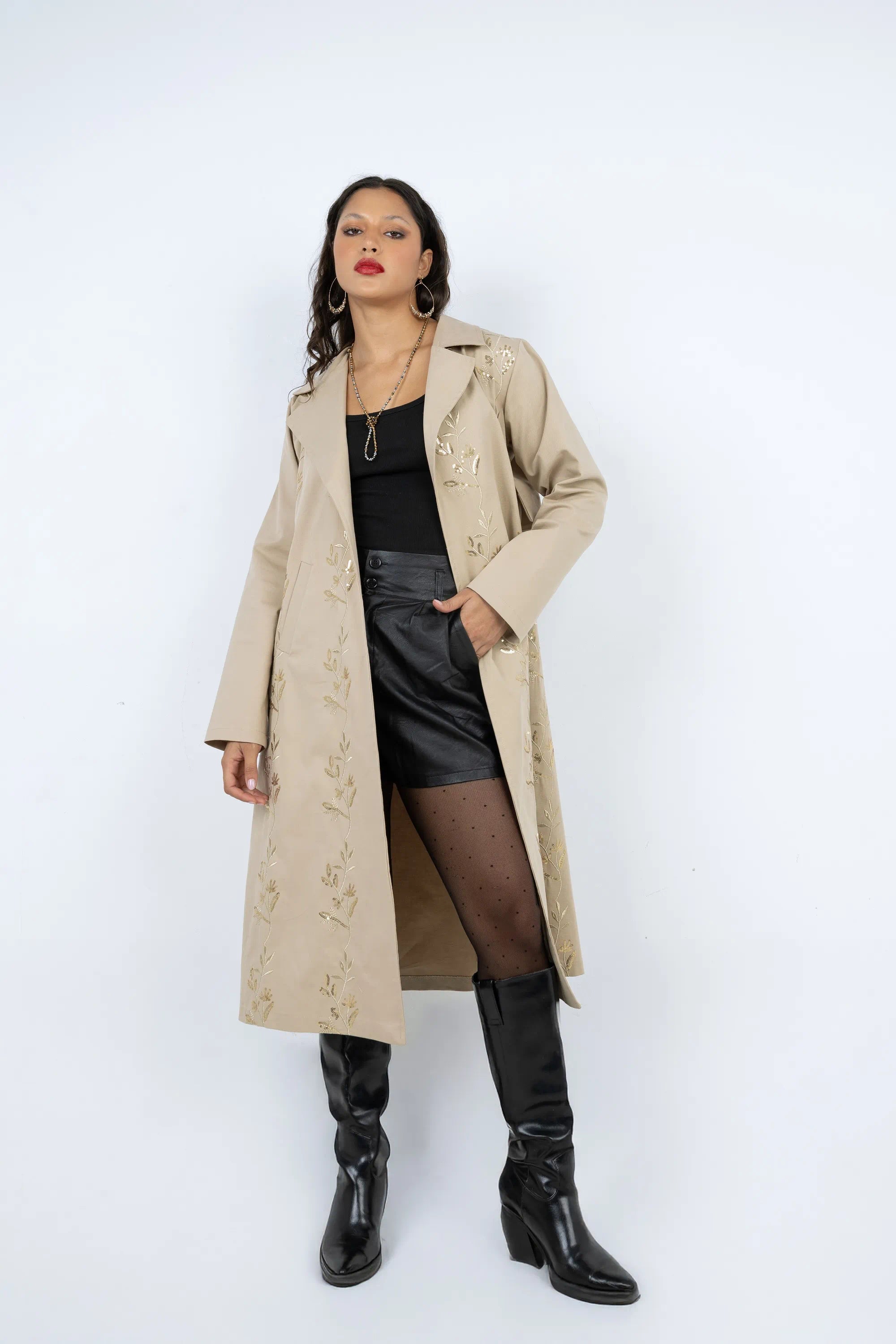 Woman wearing a beige trench coat, black top, and black knee-high boots on a white background