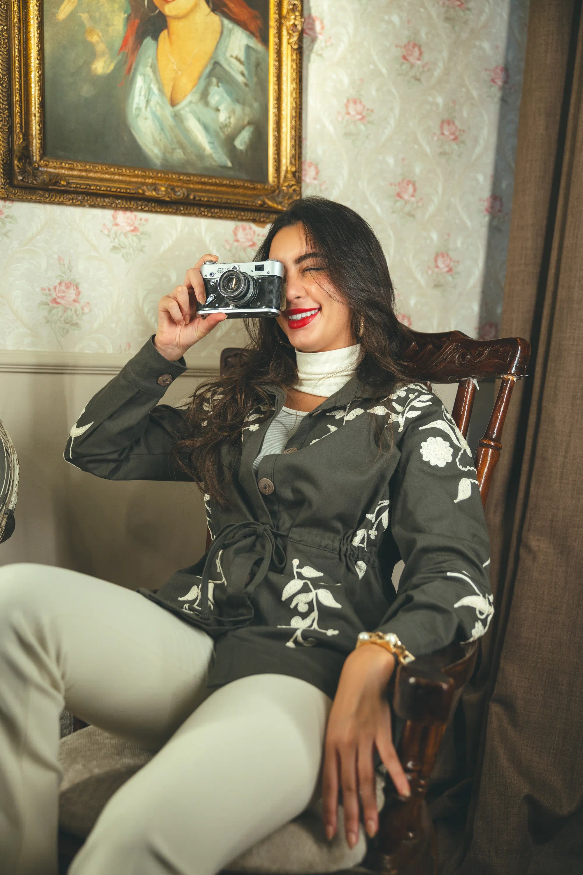 Woman taking a selfie with an old-fashioned camera in a room with floral wallpaper and a framed portrait.