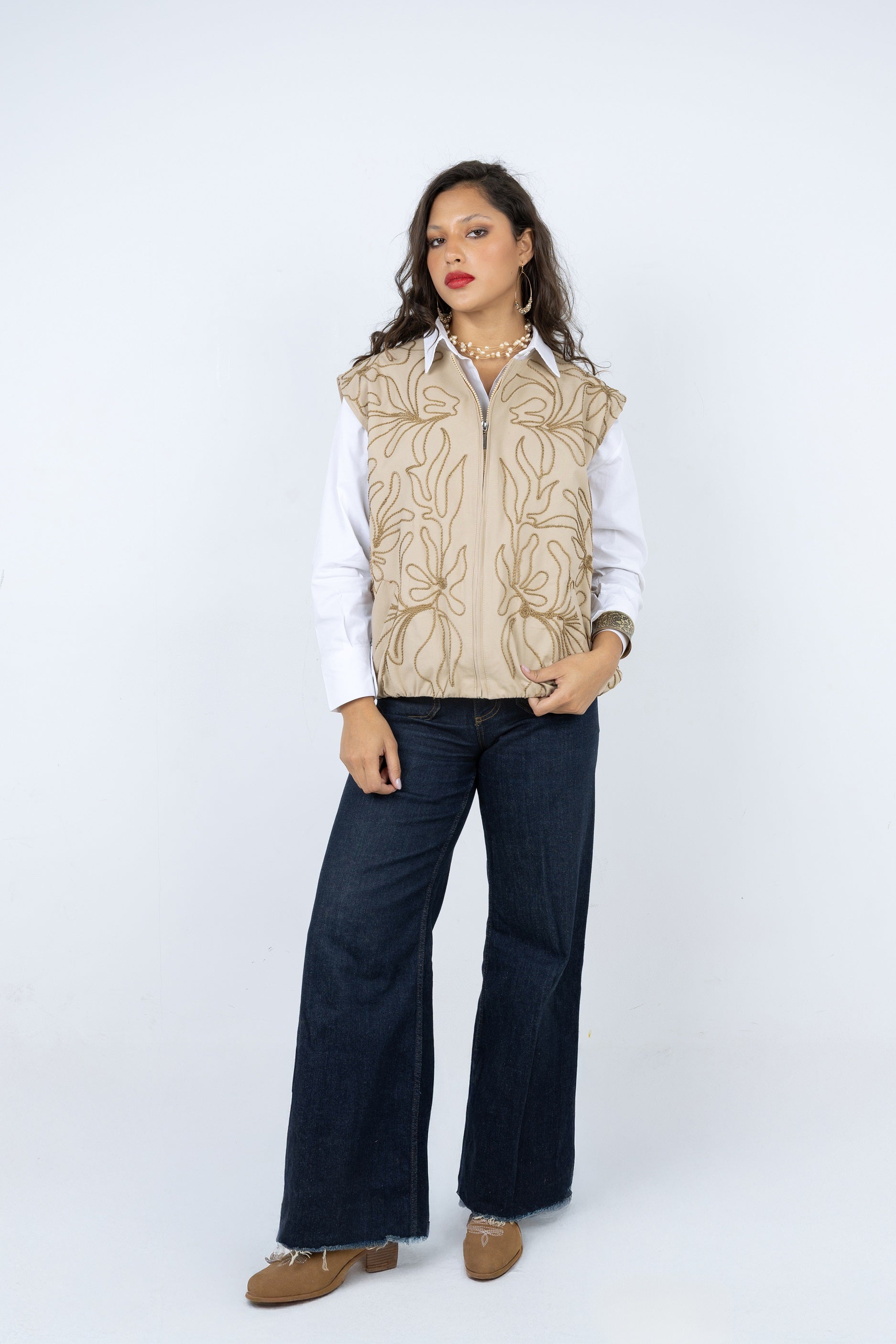 Woman wearing a beige embroidered vest over a white shirt with dark jeans on a white background