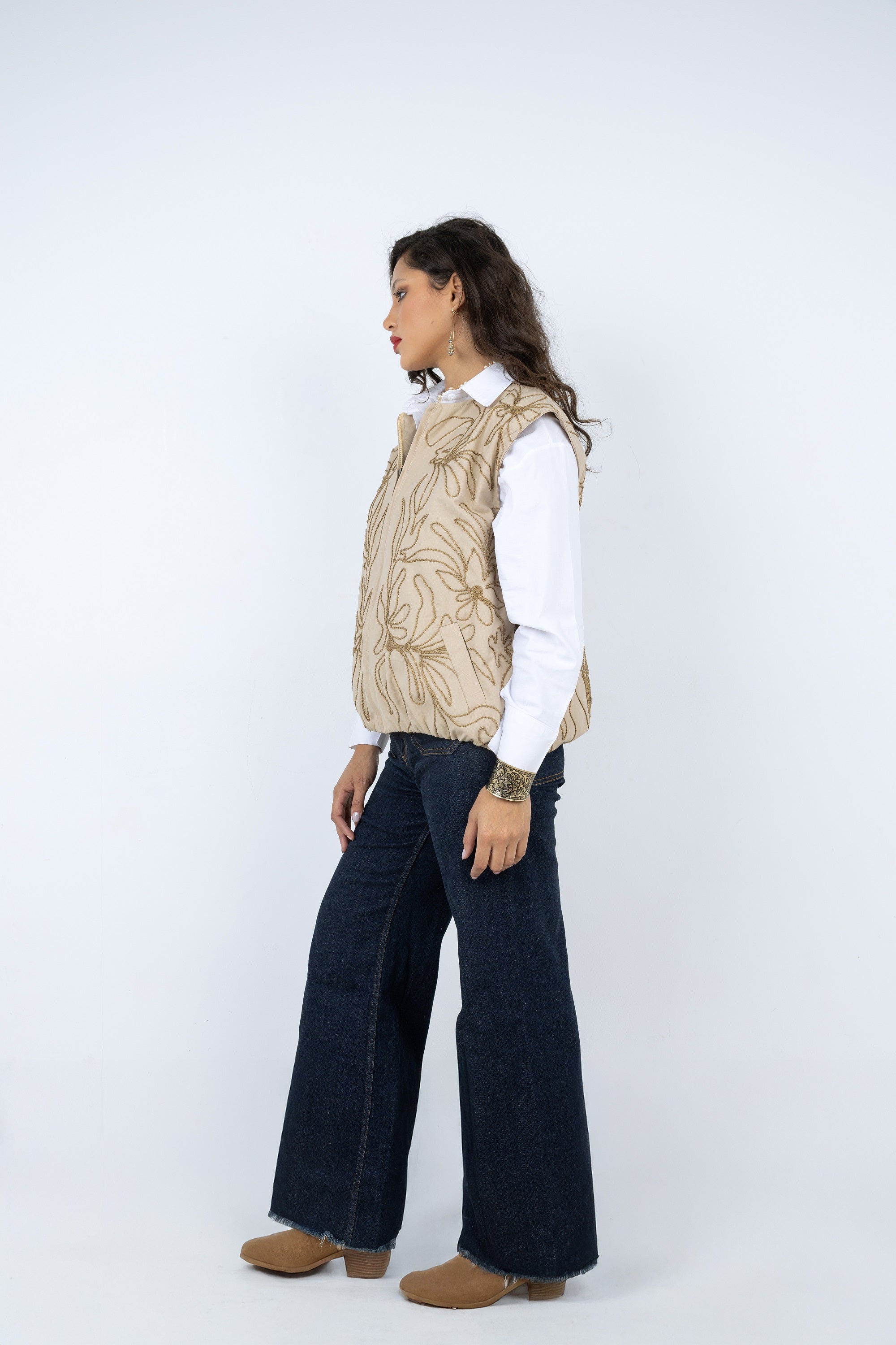 Person wearing a beige patterned vest over a white shirt with dark jeans on a white background