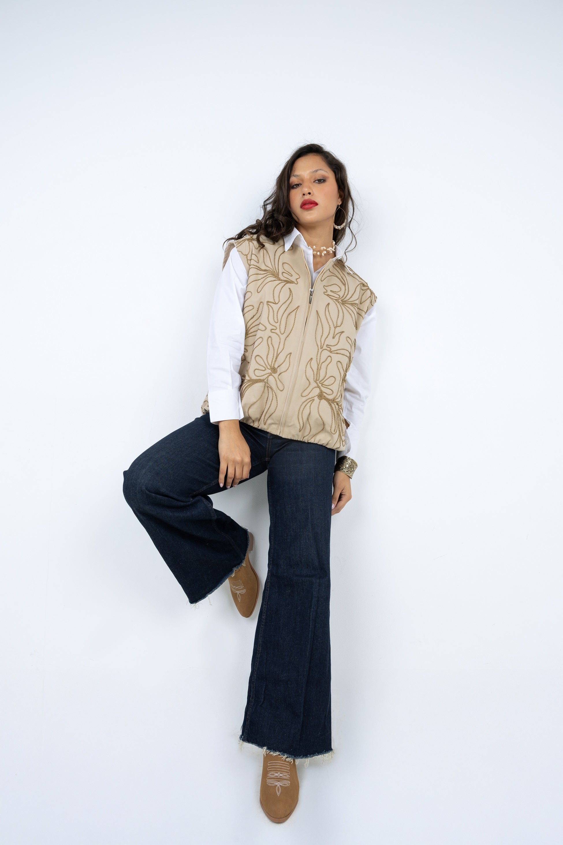 Woman wearing a beige patterned vest over a white shirt with dark blue jeans on a white background