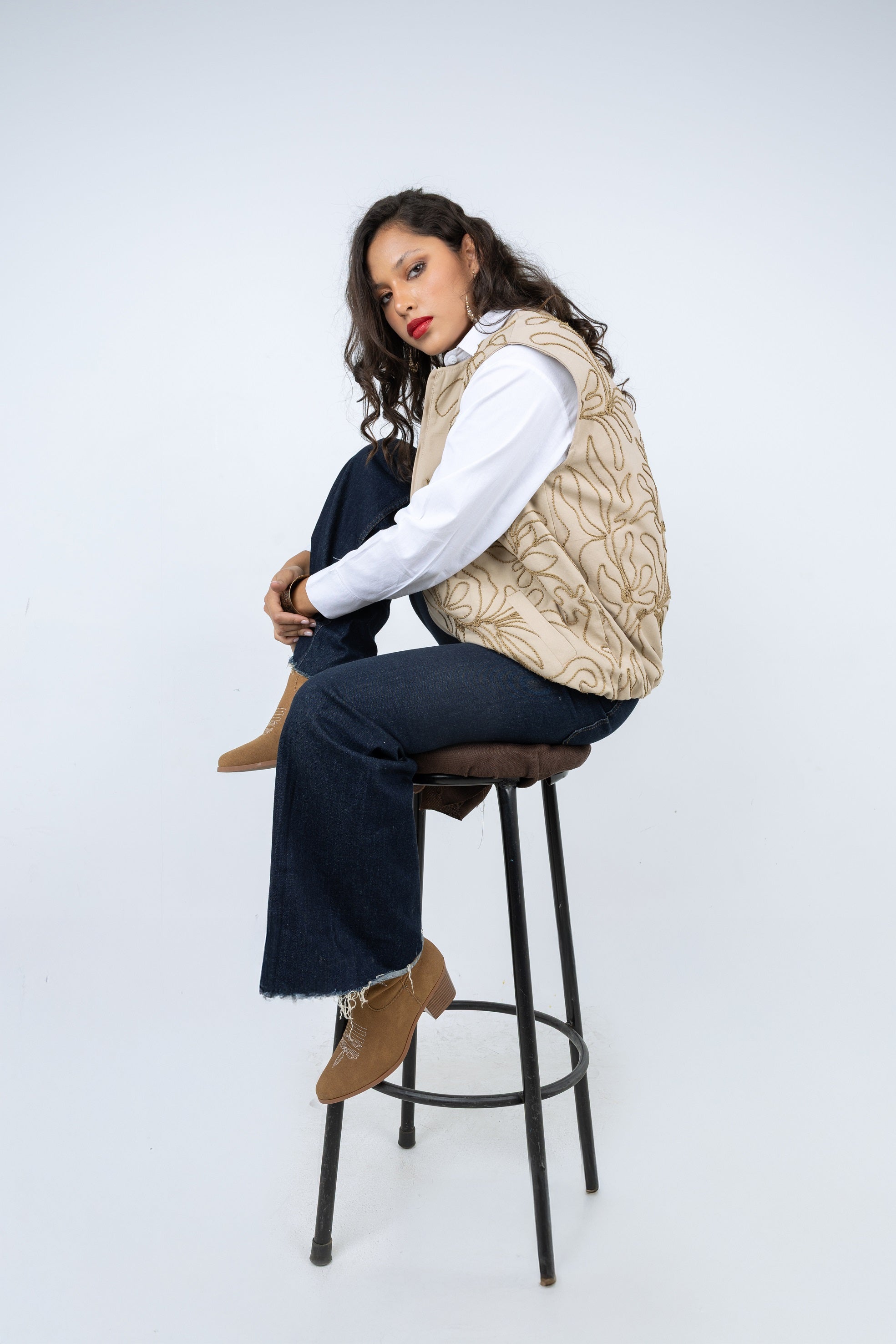 Woman sitting on a stool wearing a beige cable knit vest against a white background