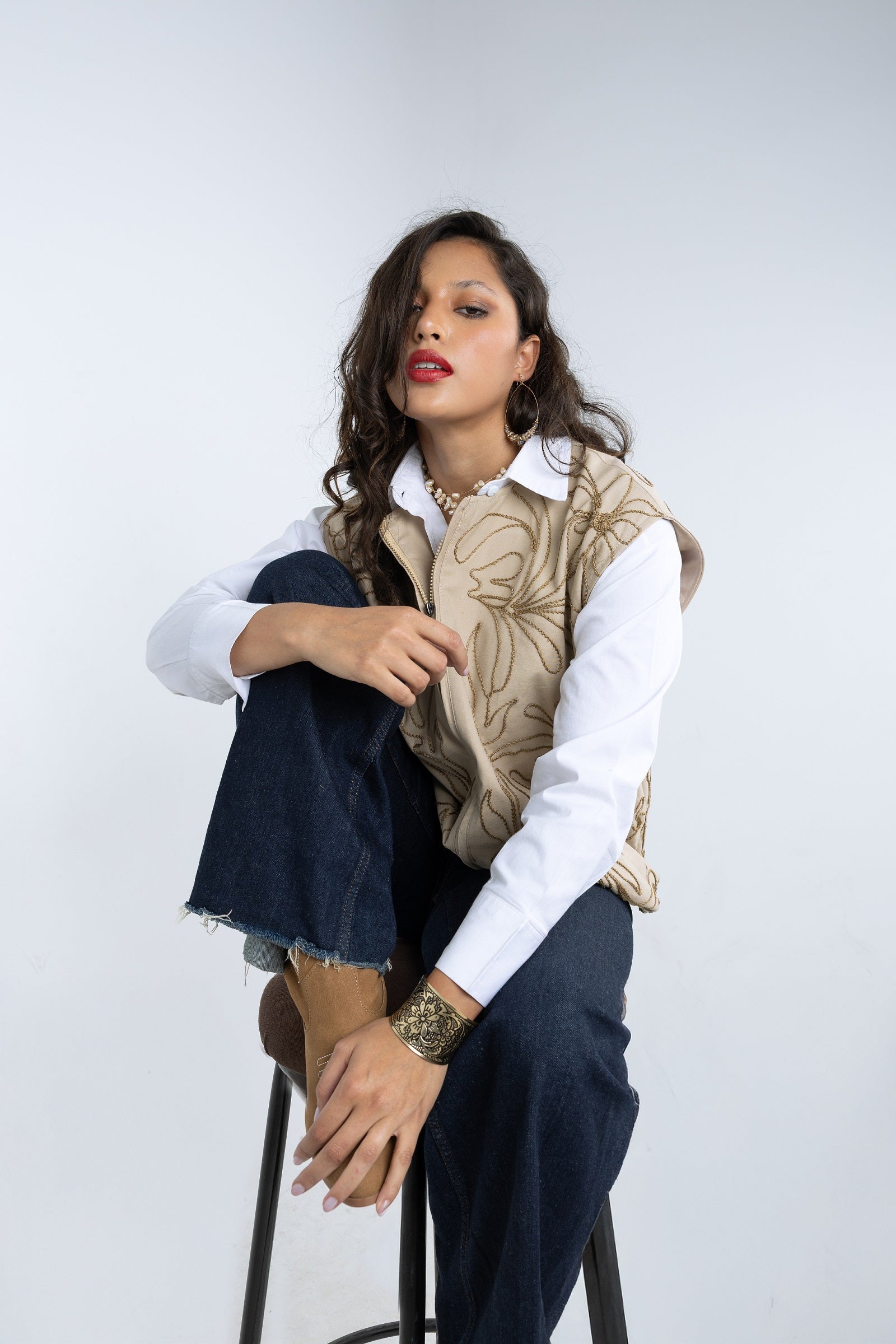 Woman sitting on a stool wearing a white shirt, dark pants, and a patterned vest against a plain background