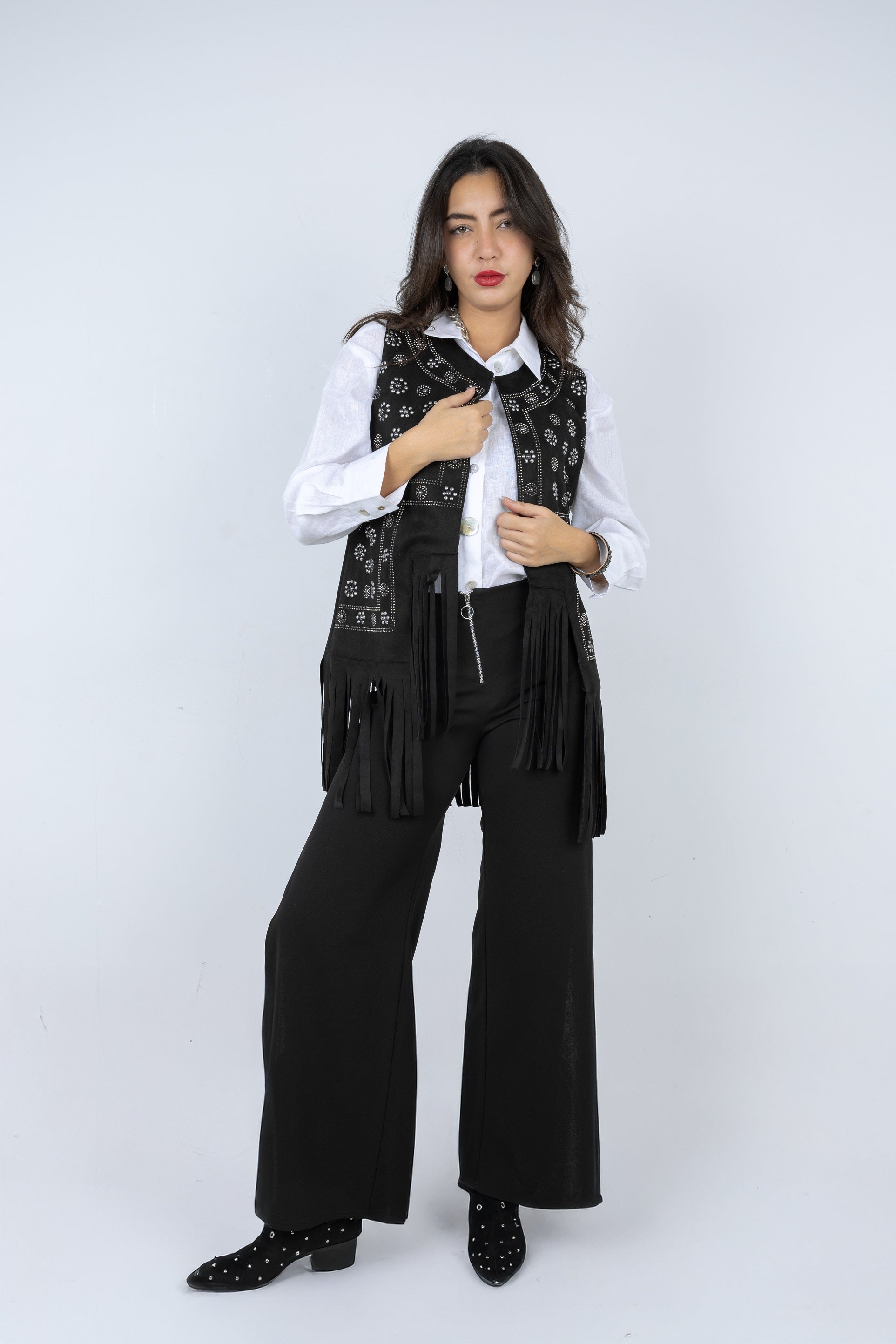 Woman wearing a black vest with fringe, white shirt, and black pants on a white background