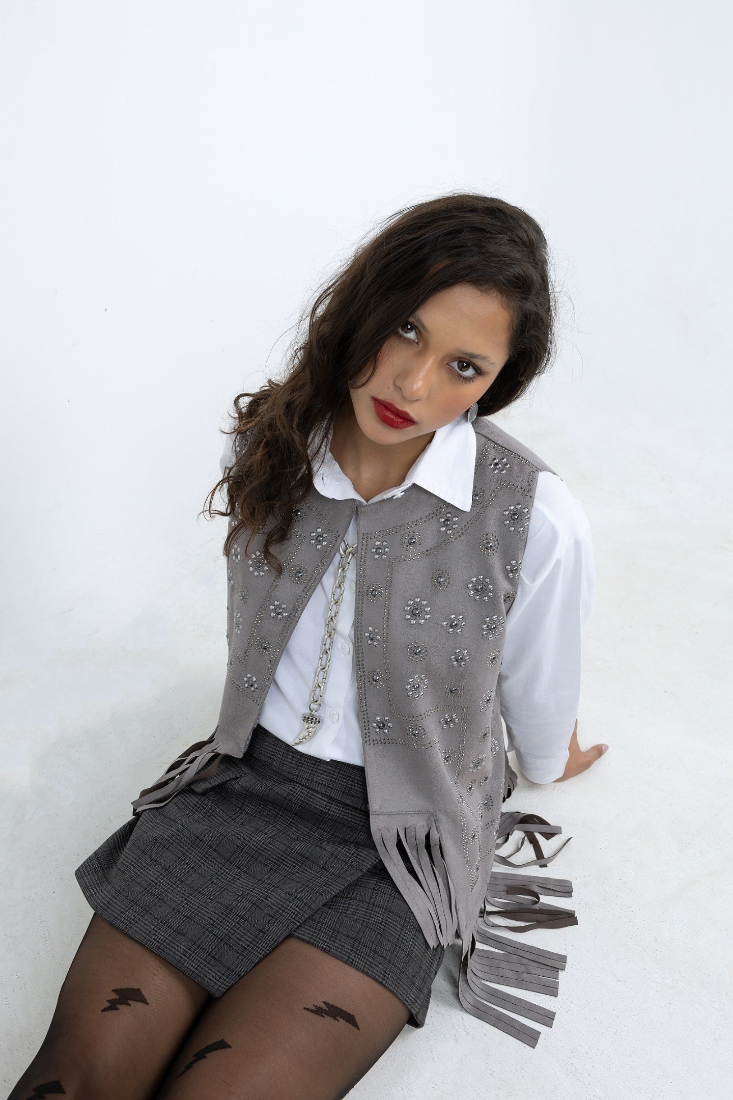 Woman wearing a gray vest with fringe, white shirt, and plaid skirt on a white background