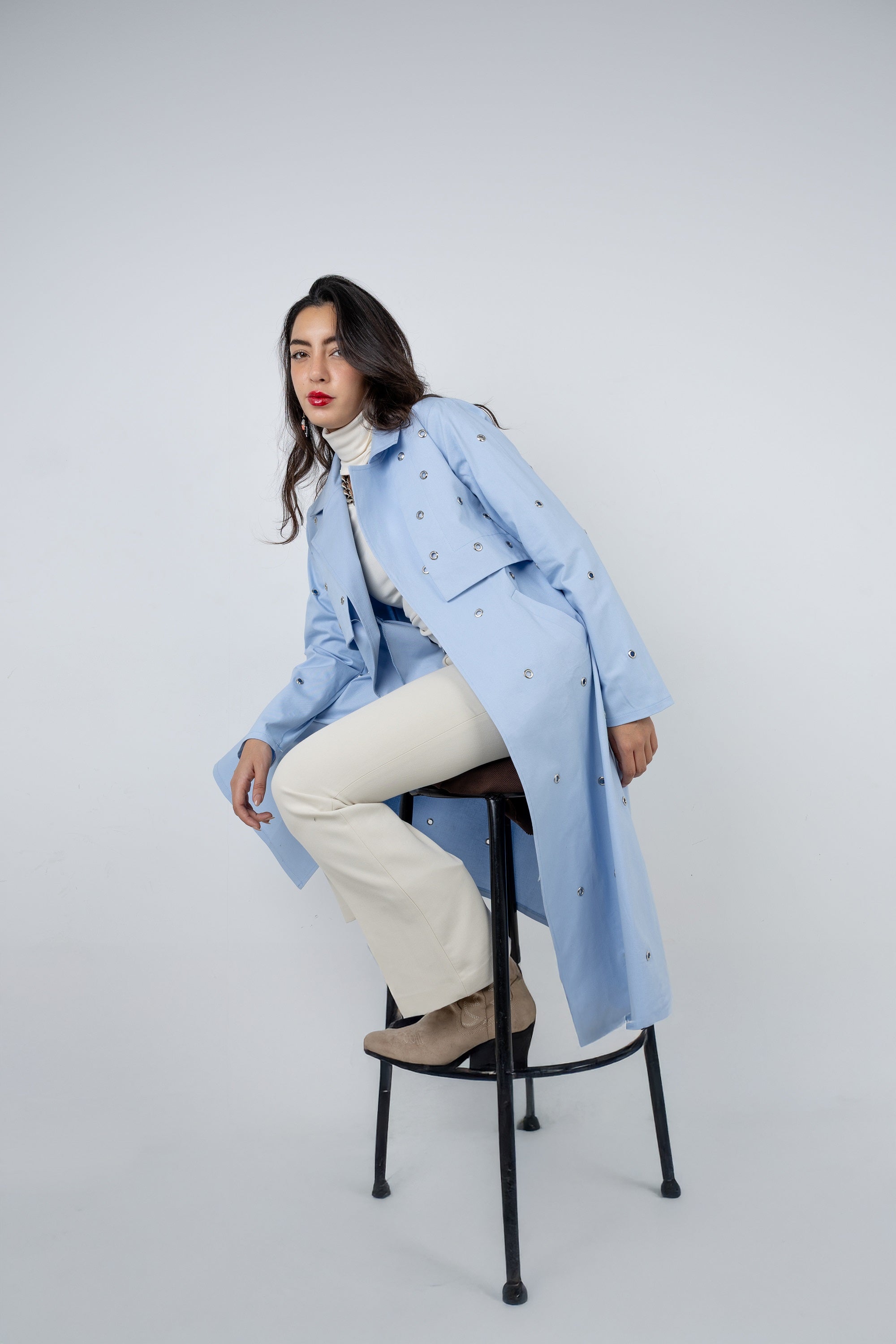 Woman wearing a light blue coat sitting on a stool against a white background