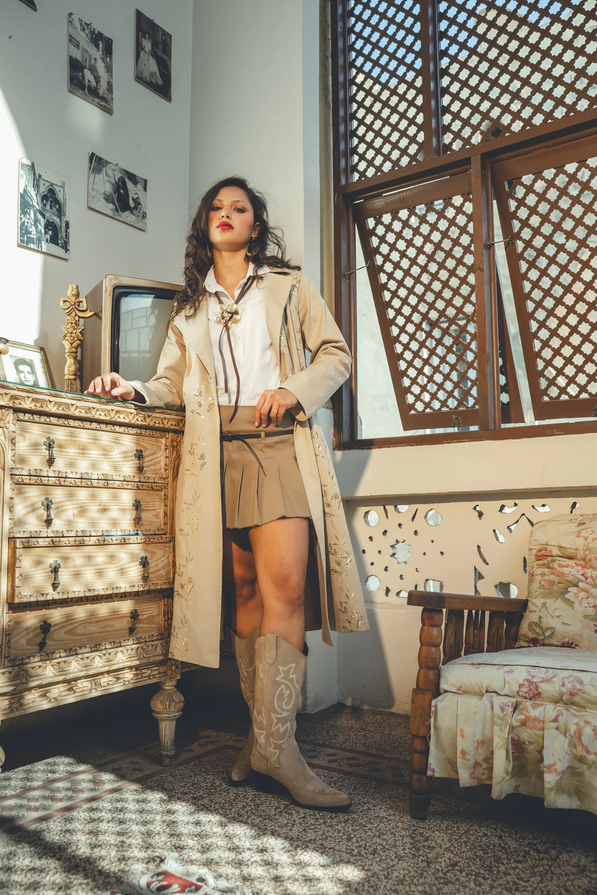 Woman in a beige coat and boots standing in a room with wooden furniture and a window with lattice design.