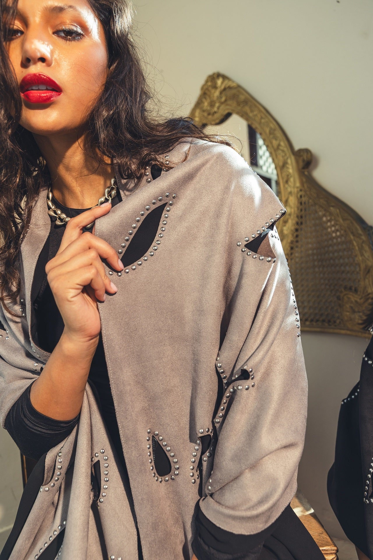 Woman wearing a beige shawl with black accents, standing in front of a decorative mirror.