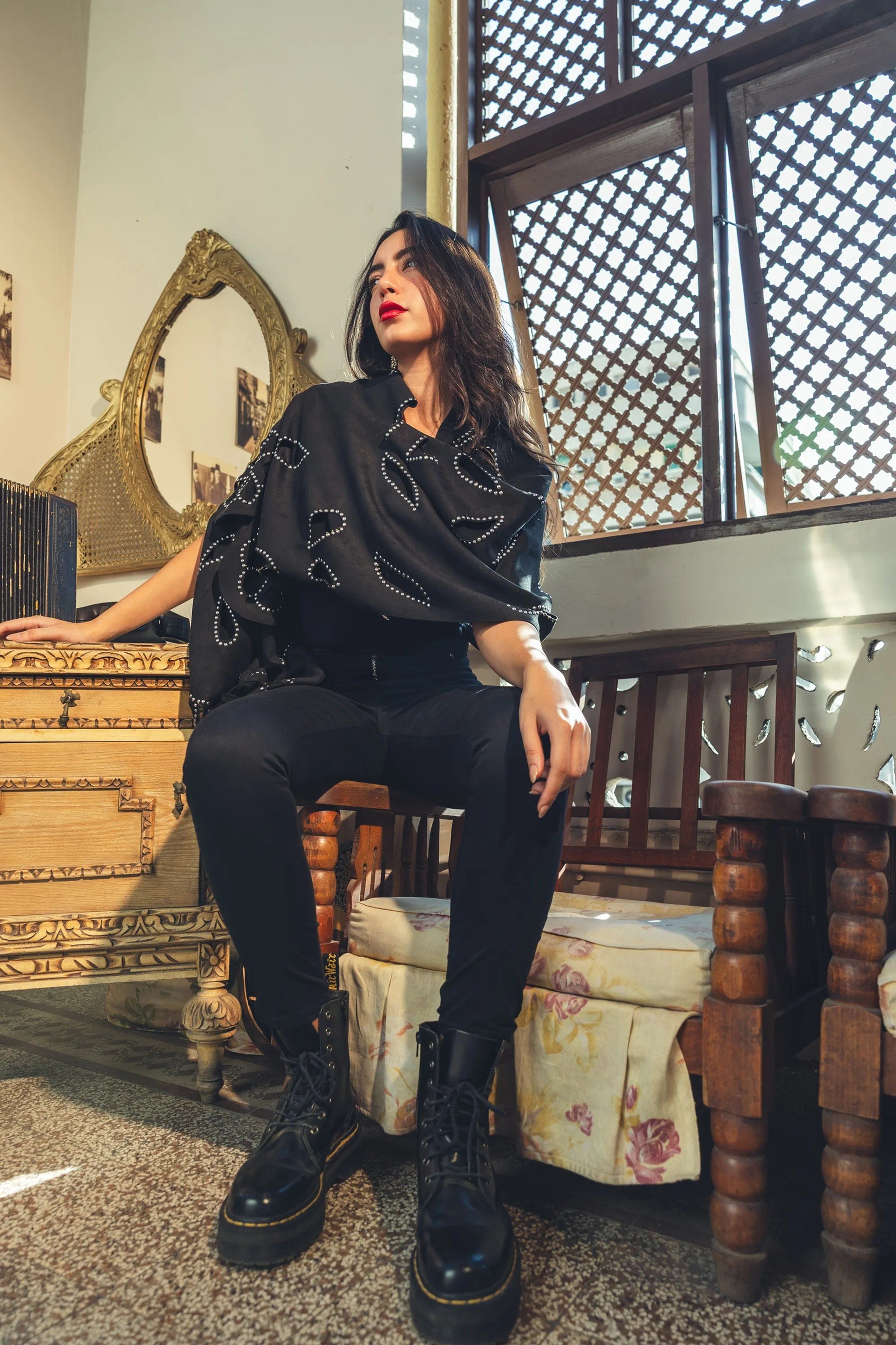 Woman in black outfit with embellishments sitting on a wooden chair in an ornate room.