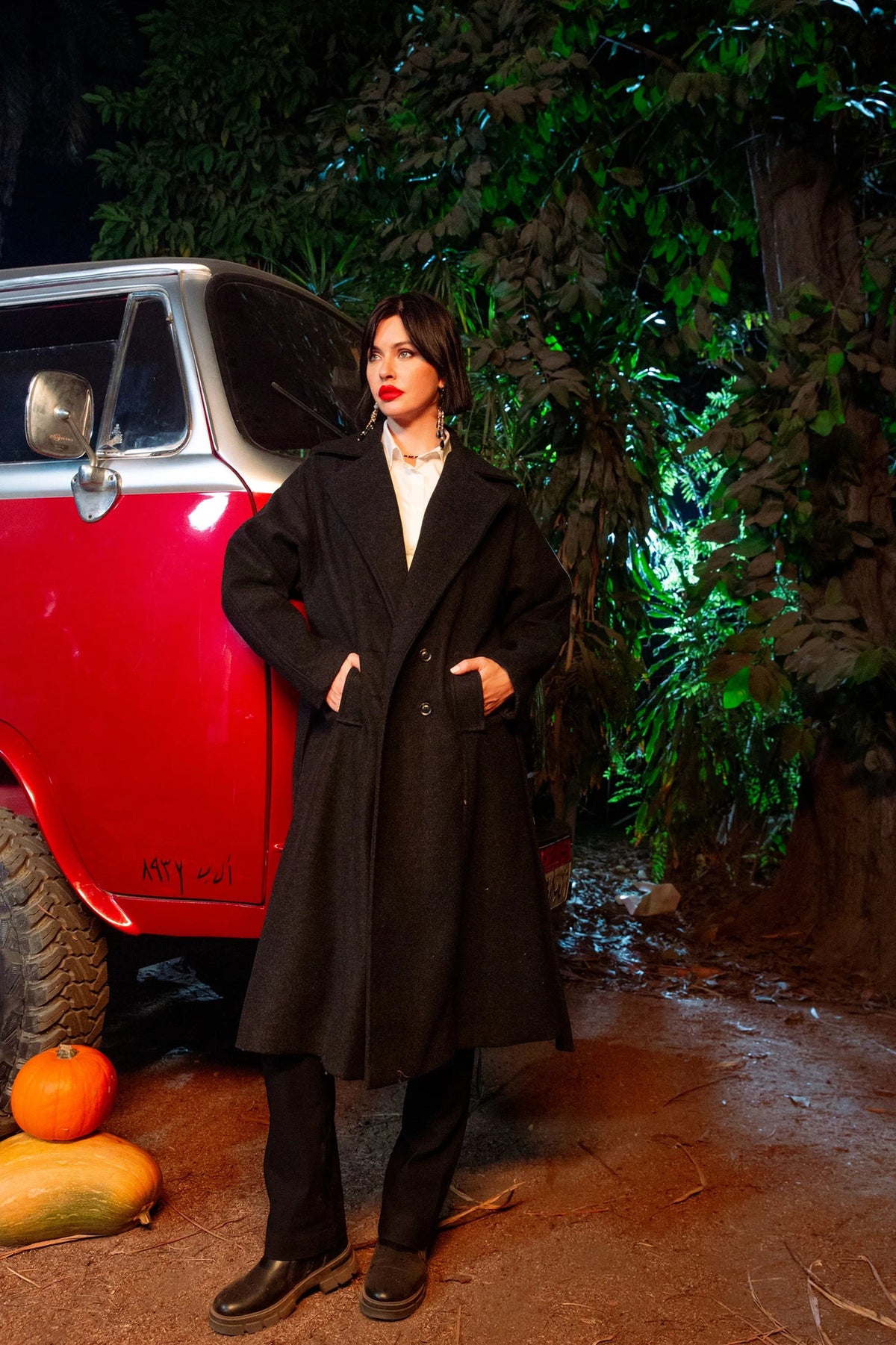 Person wearing a black coat standing next to a red vehicle with greenery in the background