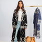Woman wearing a long black and white patterned coat with a plain white shirt and light blue jeans, standing next to a basket of pumpkins and another patterned coat on a rack.