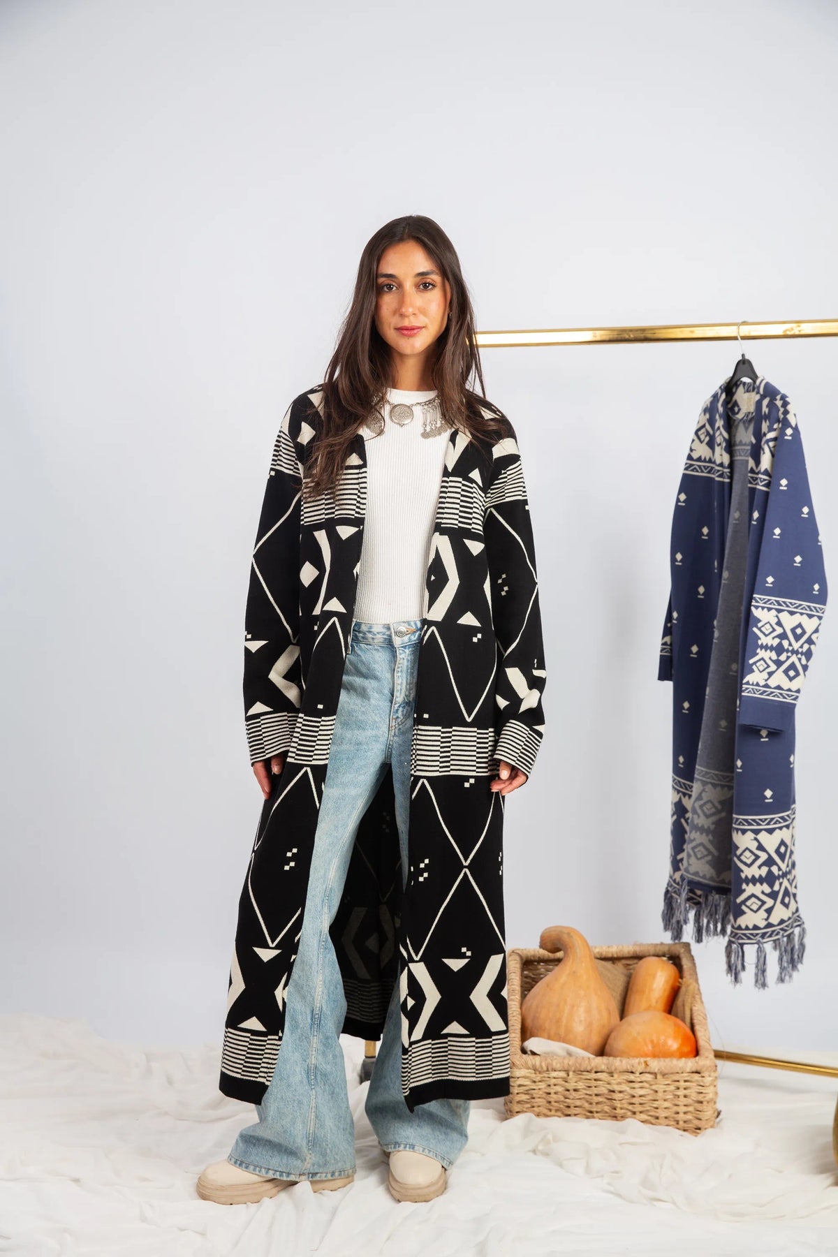 Woman wearing a long black and white patterned coat with a plain white shirt and light blue jeans, standing next to a basket of pumpkins and another patterned coat on a rack.