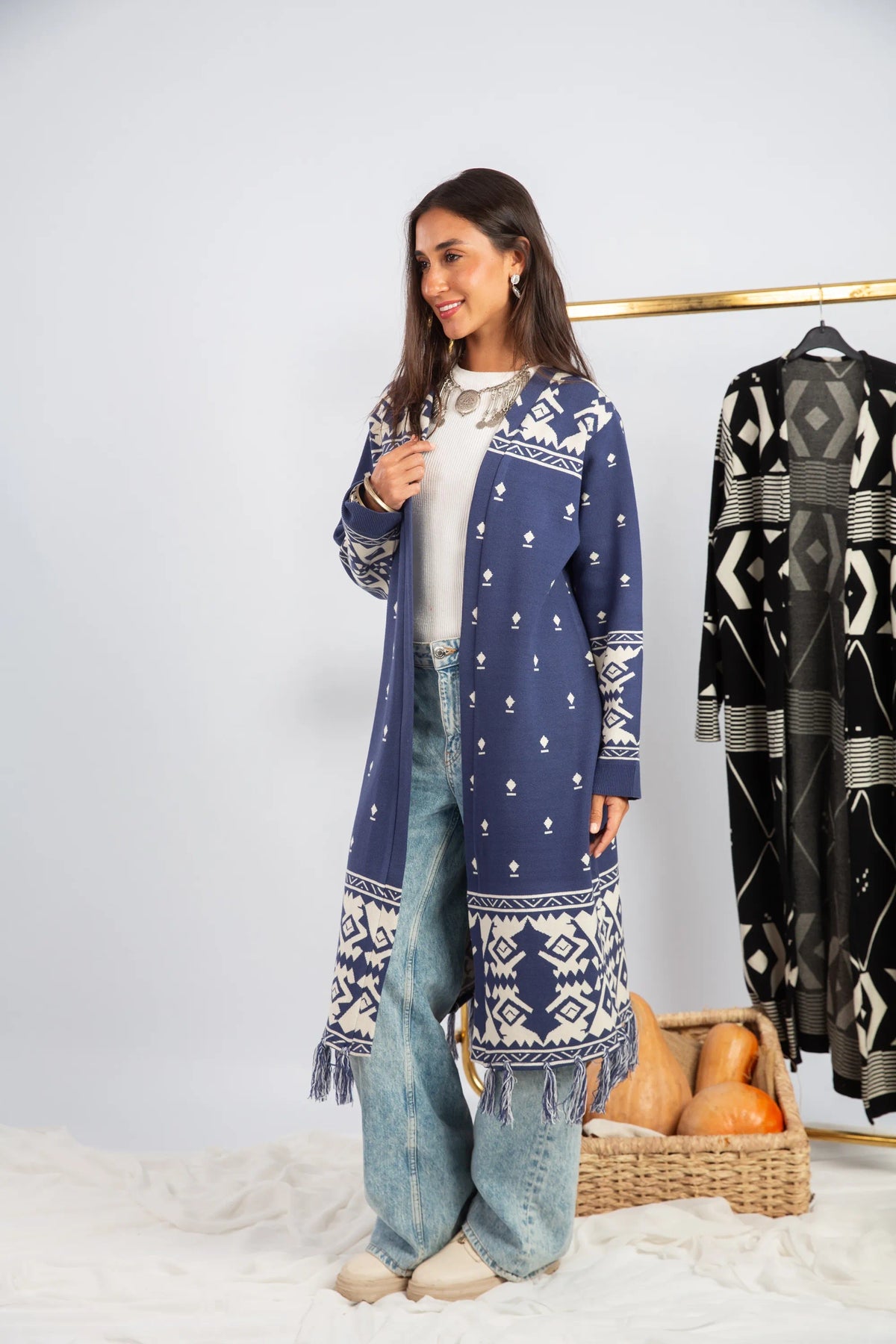 Woman wearing a long blue patterned coat with a white shirt and jeans, standing next to a basket of oranges.
