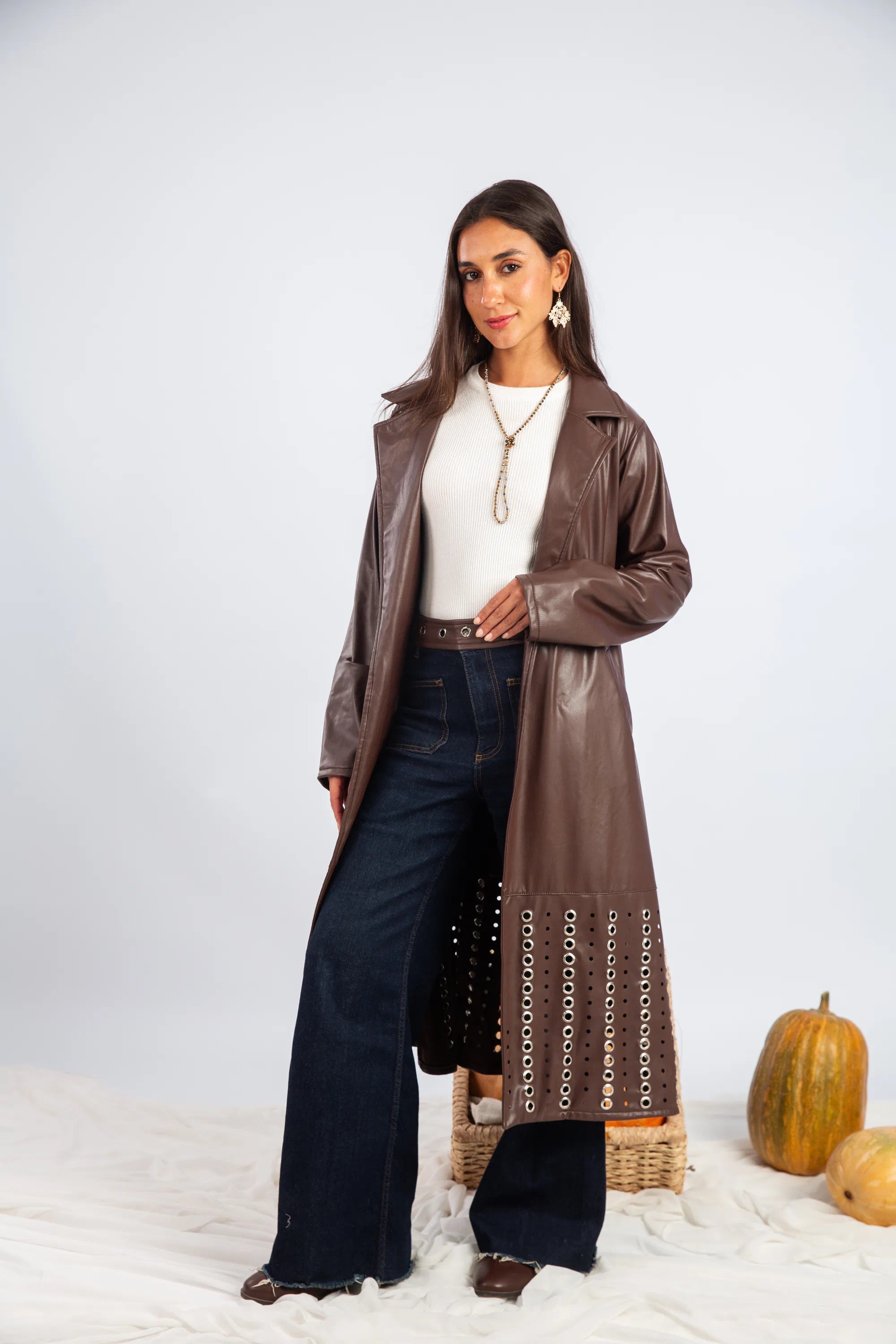 Woman wearing a long brown leather coat with decorative chain details on a white background.