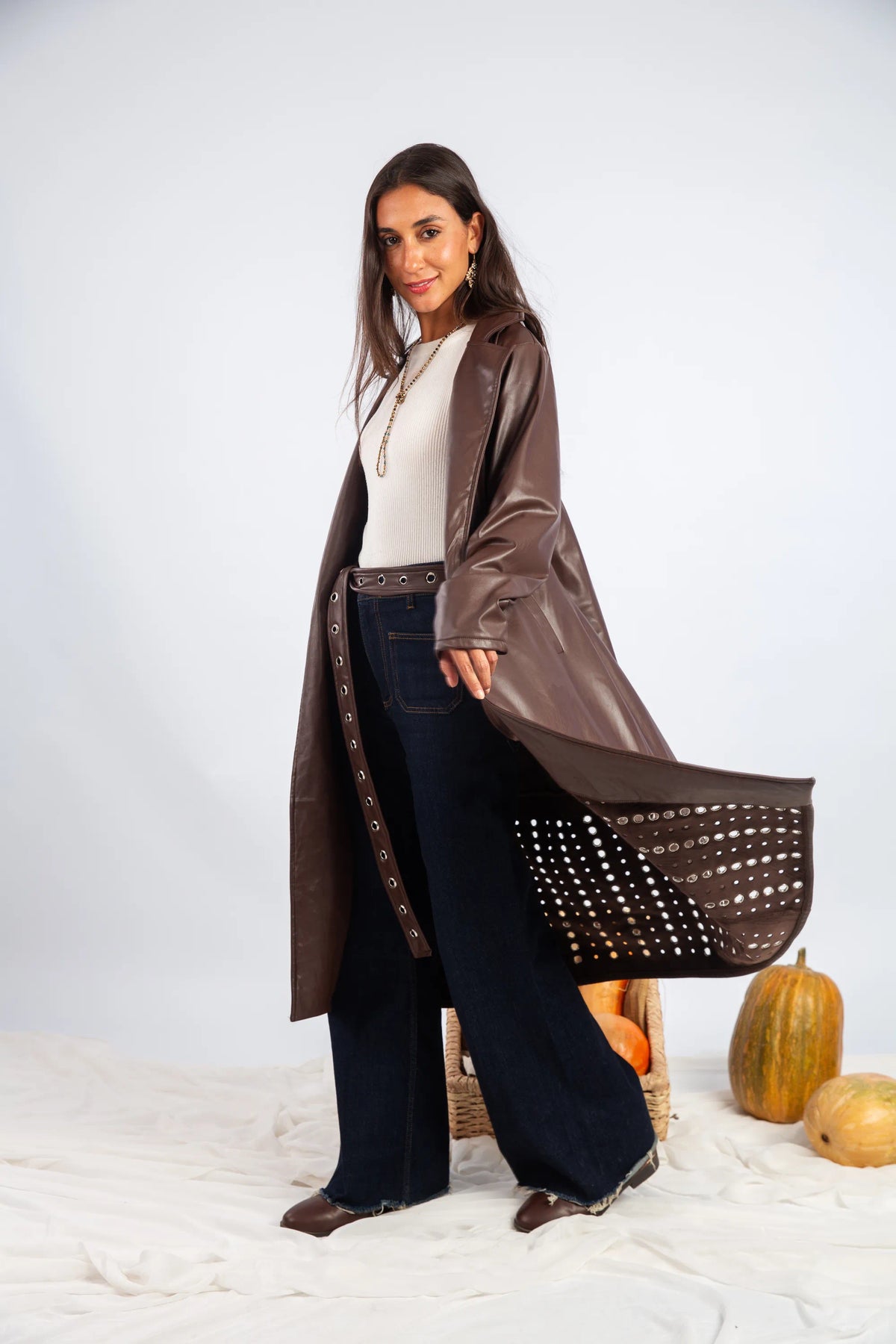 Woman wearing a long brown coat with studded details, standing against a white background.
