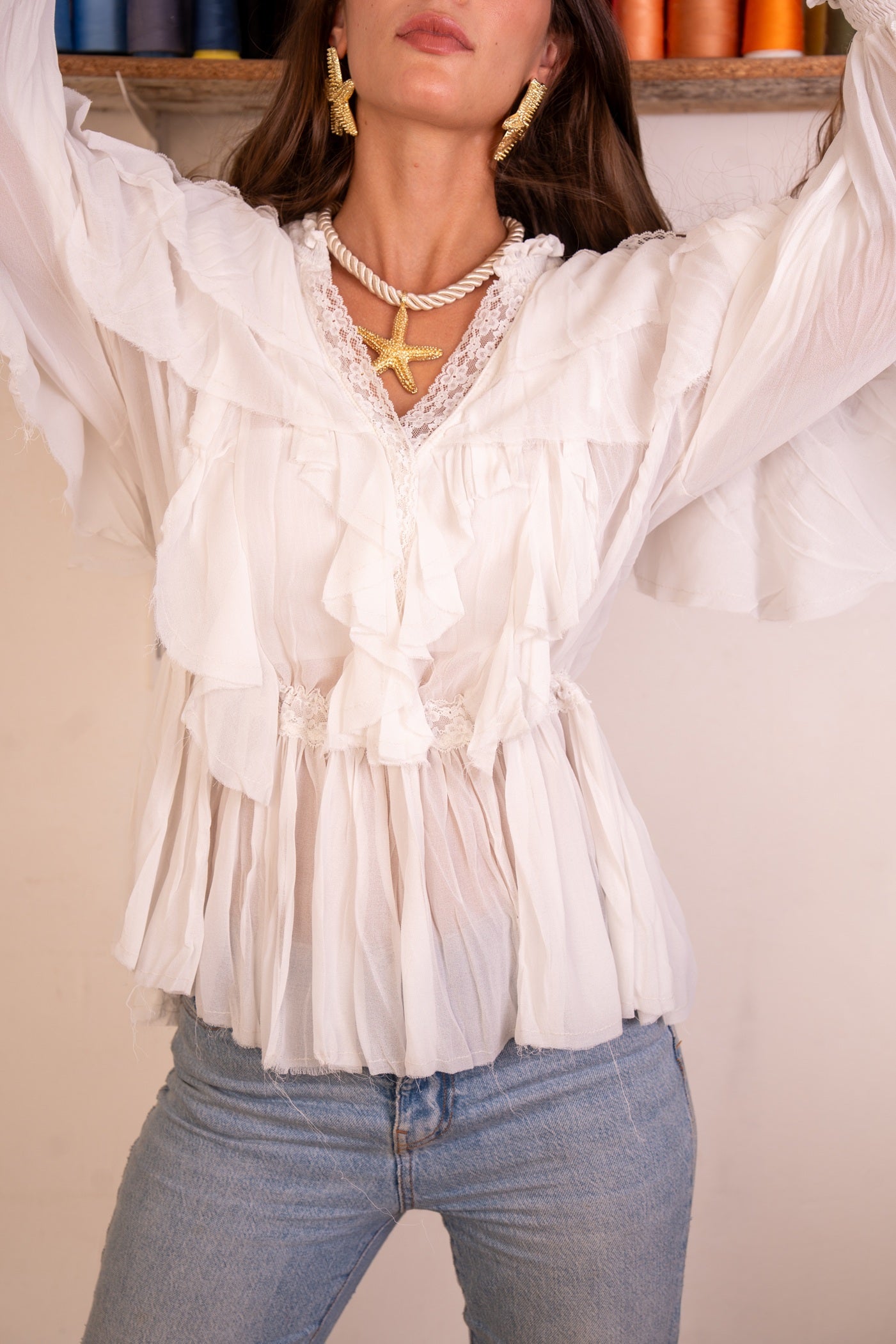 Person wearing a white blouse with ruffled details and blue jeans, standing against a neutral background.