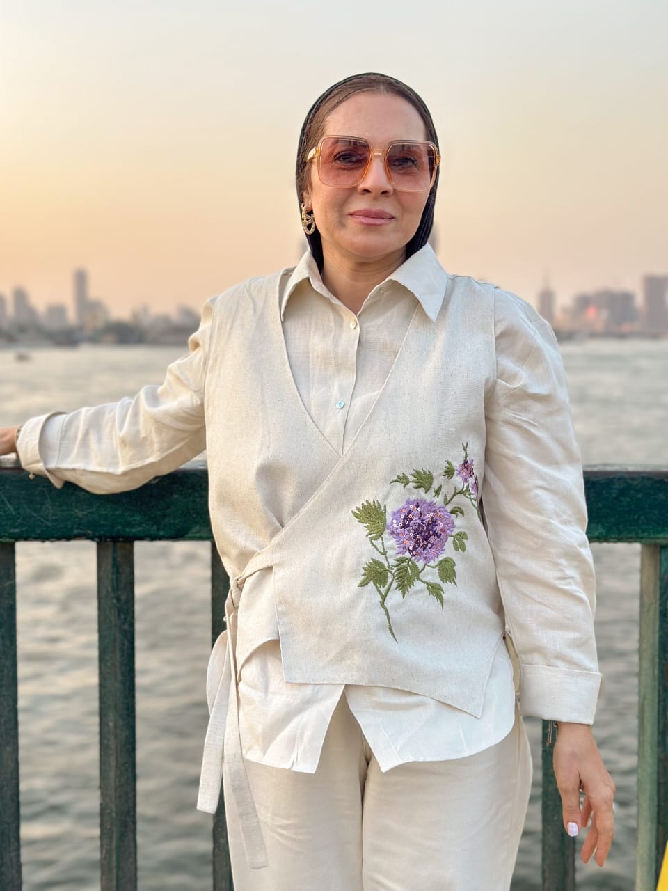 Woman in a white outfit with floral embroidery standing by a waterfront with a city skyline in the background.