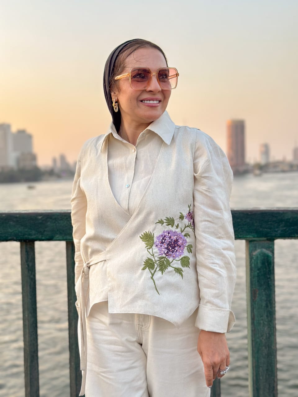 Woman in a white outfit with floral embroidery standing by a waterfront with city skyline in the background.