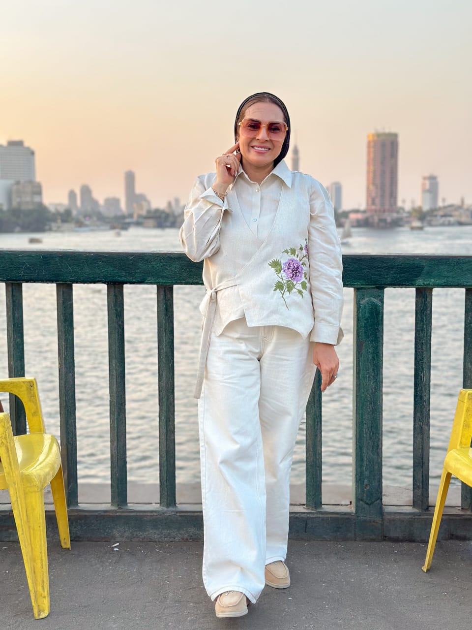 Person in a white outfit with floral embroidery standing by a waterfront with a city skyline in the background.