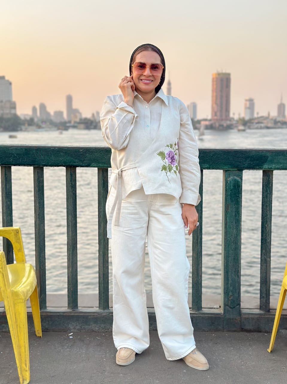 Person in a white outfit with floral details standing by a waterfront with city skyline in the background