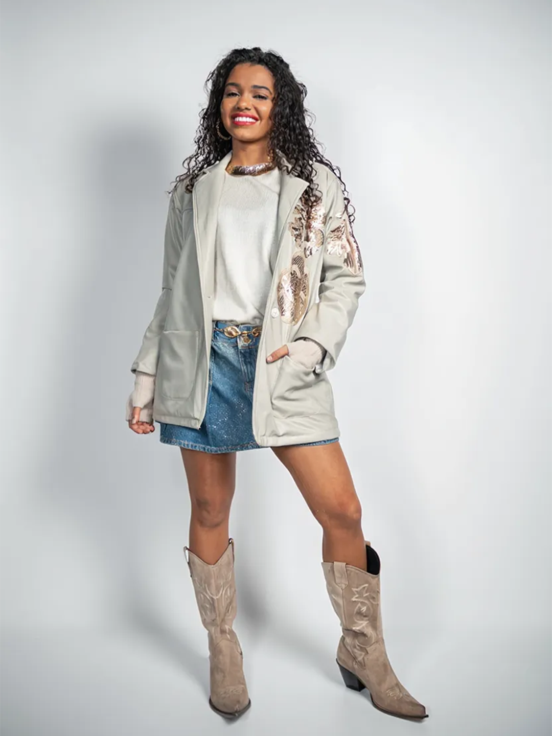 Woman wearing a light gray jacket, white shirt, denim skirt, and brown boots on a plain background