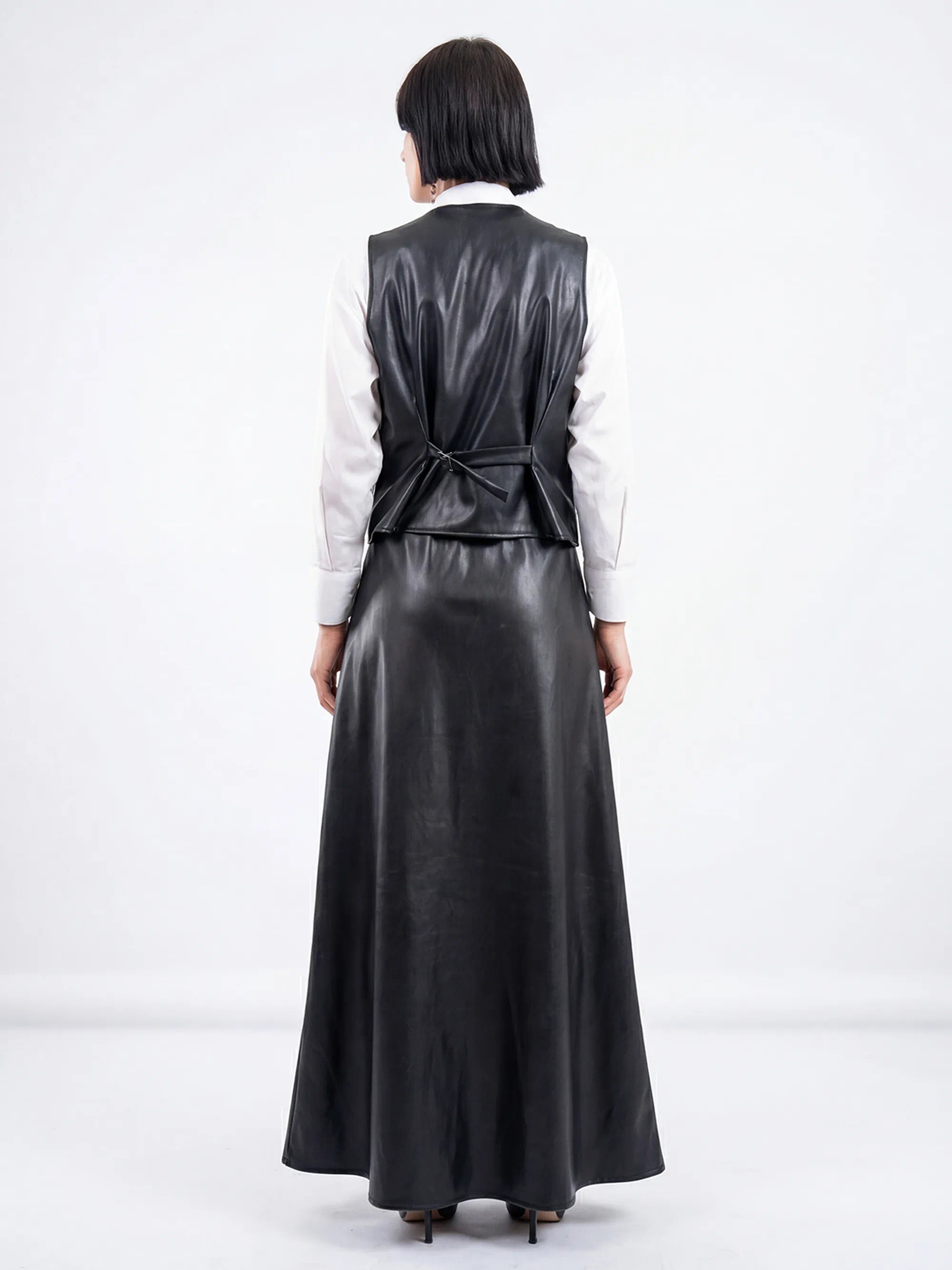 Person wearing a long black leather dress with a fitted waist and a white shirt underneath on a white background.